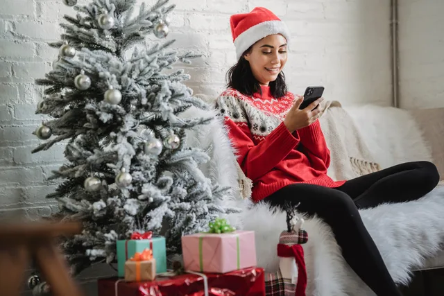 Young female in red sweater sitting on sofa near decorated Christmas tree and taking selfie on smartphone. Sådan starter du en dansk webshop